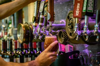 Hand holding a tulip glass as frothy amber craft beer pours from a row of draft taps with decorative gears and wine bottles in the background under purple bar lighting