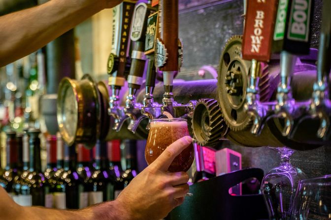 Hand holding a tulip glass as frothy amber craft beer pours from a row of draft taps with decorative gears and wine bottles in the background under purple bar lighting