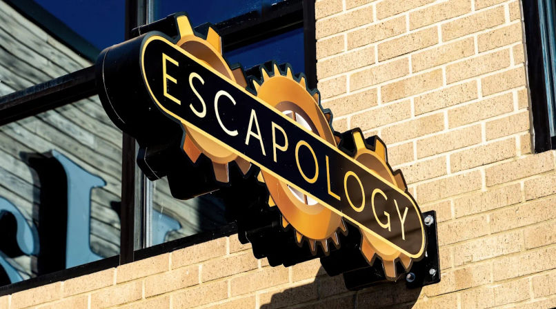 Sunlit black-and-gold interlocking-gear sign for an escape room venue mounted on a tan brick storefront