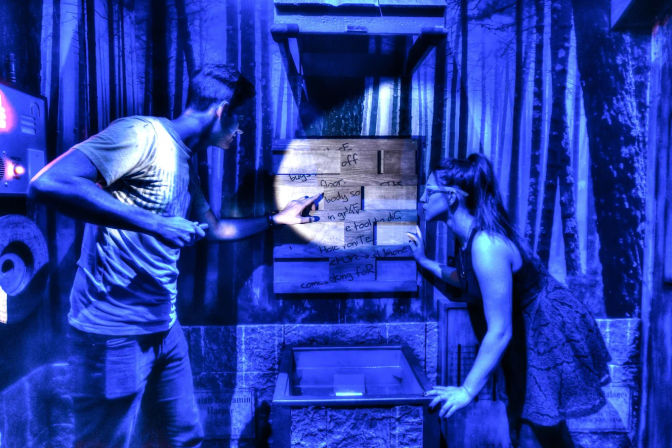 Two people in a blue-lit escape room examine a wooden clue board on a forest mural, one pointing with a flashlight.
