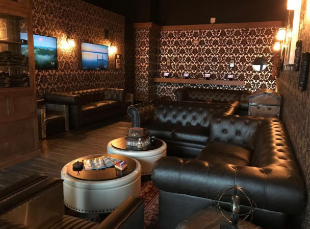 Dimly lit vintage lounge with tufted black leather Chesterfield sofas, round ottoman coffee tables, ornate damask wallpaper, warm wall sconces and wall-mounted TVs.