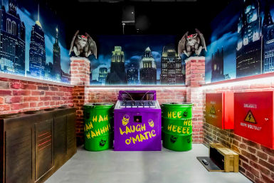 Comic-book–style themed room with faux brick walls and a nighttime city skyline mural, two gargoyle statues on pillars, a purple 'Laugh O'Matic' machine flanked by green barrels painted with manic laughter, and red high-voltage boxes.