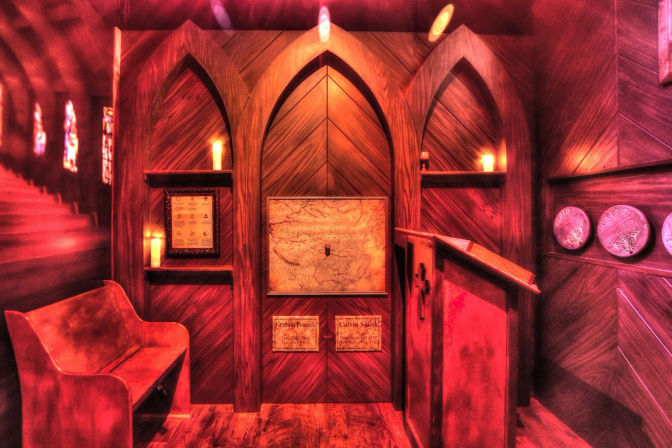 Cozy, warm-lit wooden chapel alcove with three gothic arched panels, a framed map and memorial plaques, flickering candles on shelves, a wooden bench and lectern, and blurred stained-glass windows.