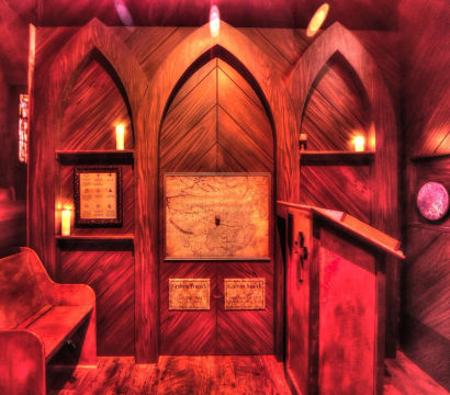 Cozy, warm-lit wooden chapel alcove with three gothic arched panels, a framed map and memorial plaques, flickering candles on shelves, a wooden bench and lectern, and blurred stained-glass windows.