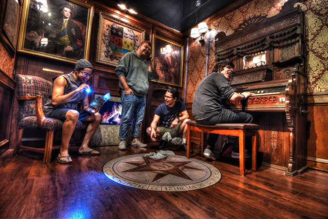Four people in a Victorian-style parlor with ornate wallpaper and oil portraits — one playing a carved pump organ, another reading by flashlight, a third on a phone, and a compass star inlaid on the wooden floor.