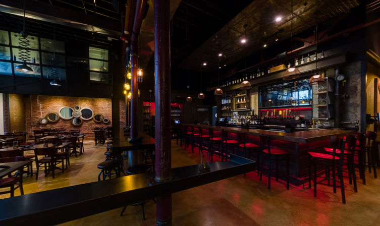 Cozy industrial-style bar interior with exposed brick, decorative round mirrors, long wooden bar with red stools, pendant lighting and empty dining tables under warm ambient glow.