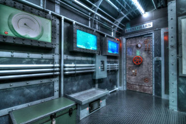 Submarine-style industrial interior with riveted steel walls, exposed pipes, twin blue underwater monitors, and a heavy porthole hatch door with a red wheel.