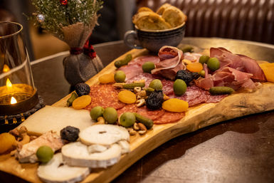 Cozy holiday charcuterie board on a wooden board with sliced salami and prosciutto, assorted cheeses, green olives, dried apricots, cornichons and crackers beside a lit candle in a warm bistro setting.