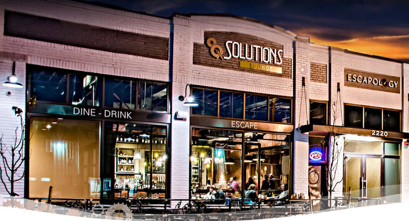 Evening downtown storefront with illuminated lounge and escape-room entrance, large glass windows revealing a lively bar and seated patrons, outdoor seating and neon open sign.