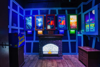 Neon-lit Scooby-themed mystery room with glowing framed paintings above a faux stone fireplace, a 'Scooby Snacks' box, wooden floor and a cartoon van window on blue-purple walls.