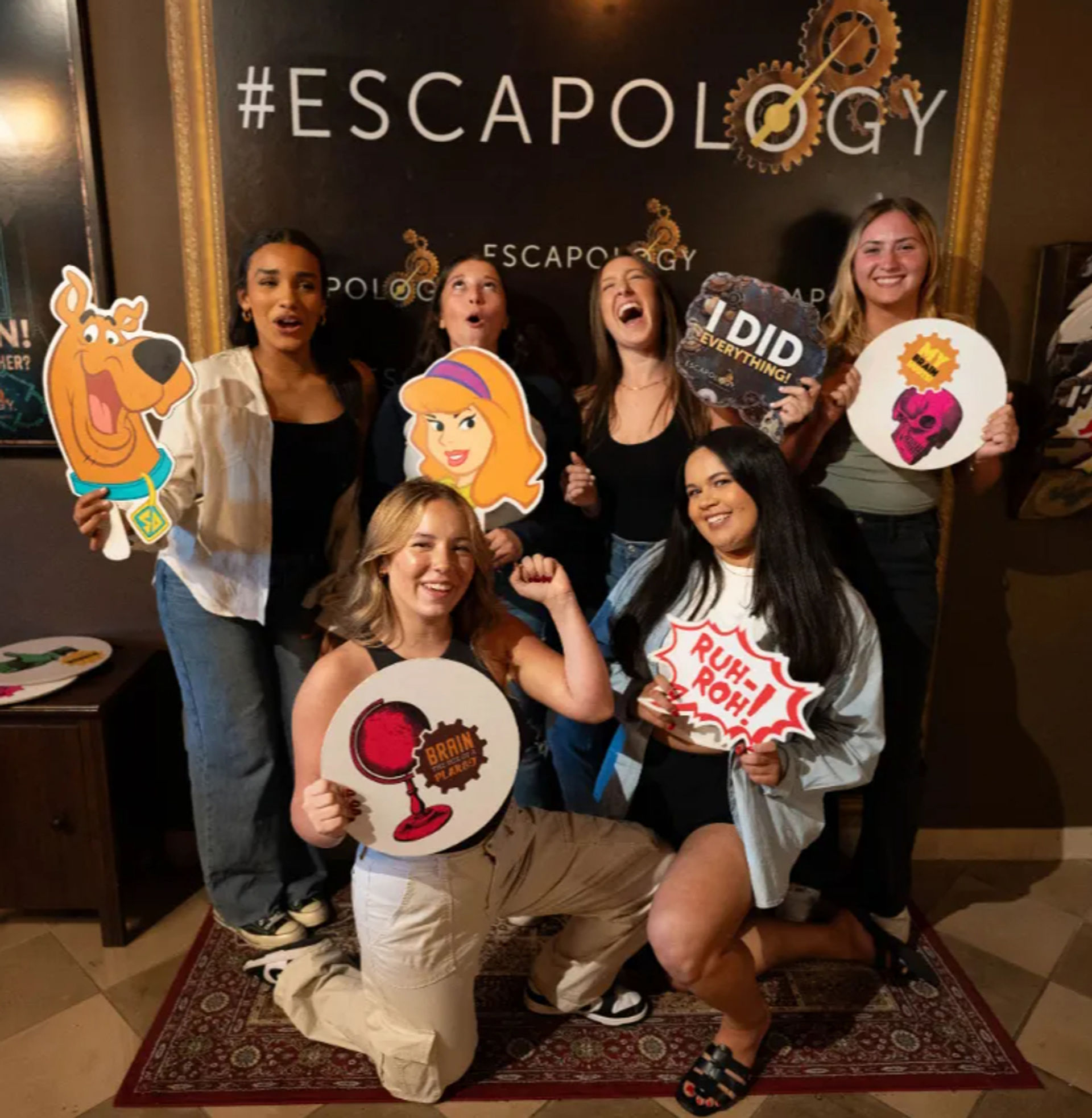Six friends pose at an indoor escape-room photo wall holding playful props — Scooby-Doo cutout, cartoon blonde, signs reading 'I DID EVERYTHING', 'RUH-ROH' and a brain trophy, celebrating a successful escape.