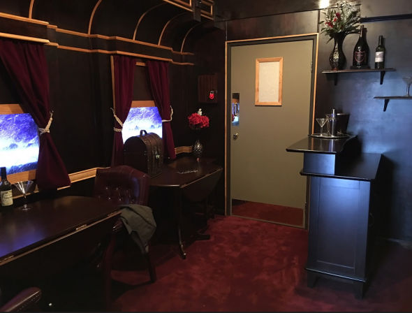 Cozy vintage-style private lounge with dark wood paneling, burgundy carpet, tufted leather chair, curtained porthole windows, a small bar counter with wine bottles and martini glasses — a retro speakeasy vibe.