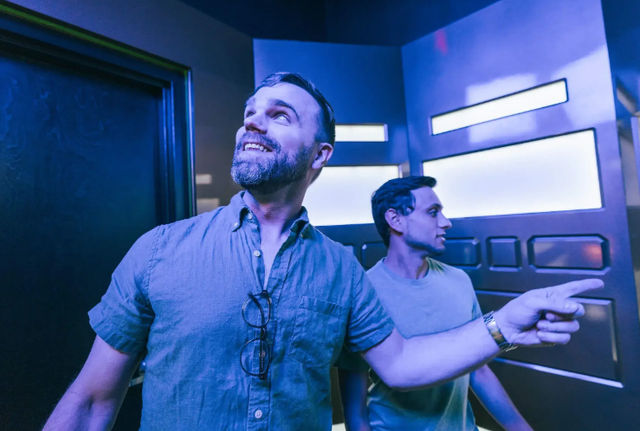 Two men in a blue neon-lit futuristic room — one pointing and smiling while the other looks up, casual shirts and glasses hanging from a collar.