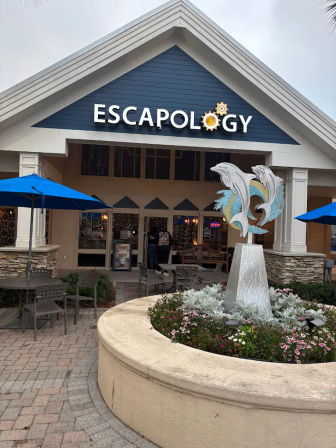 Entrance to an escape-room venue with a navy gabled sign, outdoor tables and blue umbrellas, a playful dolphin sculpture fountain in a round flowerbed, and glass double doors under an overcast sky.