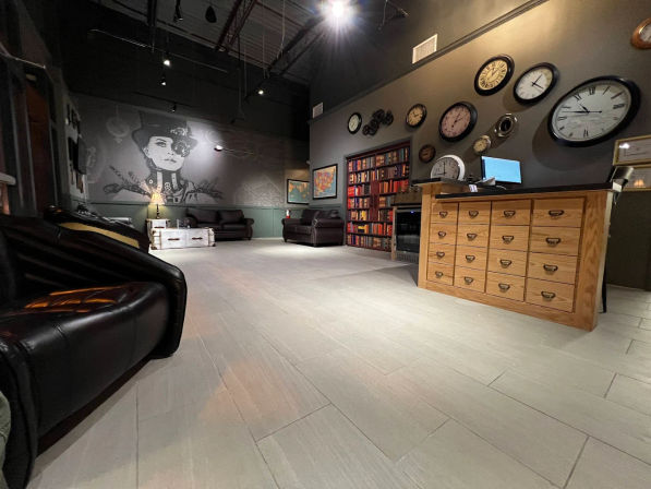 Loft-style lounge interior with light gray tile floor, black leather sofas, quirky steampunk mural of a woman, built-in bookshelf, wall of mixed vintage clocks, and a wooden reception counter with drawers.