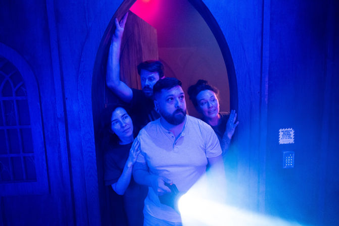 Four adults peering through a gothic arched wooden doorway into a dark room, a man in front holding a bright flashlight beam, dramatic blue-purple lighting suggesting a haunted-house or escape-room experience.