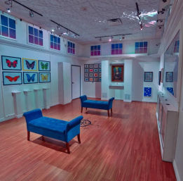 Bright modern indoor art gallery with white walls and wood floors, two blue upholstered benches, colorful framed butterfly prints on the left wall, a grid of small artworks and a framed portrait in a recessed alcove, track lighting and decorative upper windows.