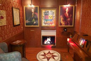 Cozy Victorian-style parlor interior with red damask wallpaper and wood paneling, glowing fireplace, three framed portraits and a coat of arms, compass floor medallion with an 'M', vintage rotary phone, piano and upholstered chair.