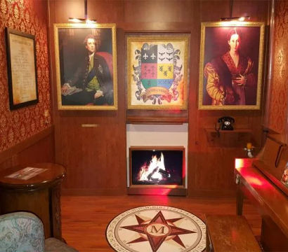 Cozy Victorian-style parlor interior with red damask wallpaper and wood paneling, glowing fireplace, three framed portraits and a coat of arms, compass floor medallion with an 'M', vintage rotary phone, piano and upholstered chair.