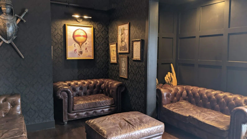 Cozy vintage lounge with dark paneled walls, two brown leather Chesterfield sofas and a matching ottoman, framed artwork including a hot-air balloon print and knight armor decor.