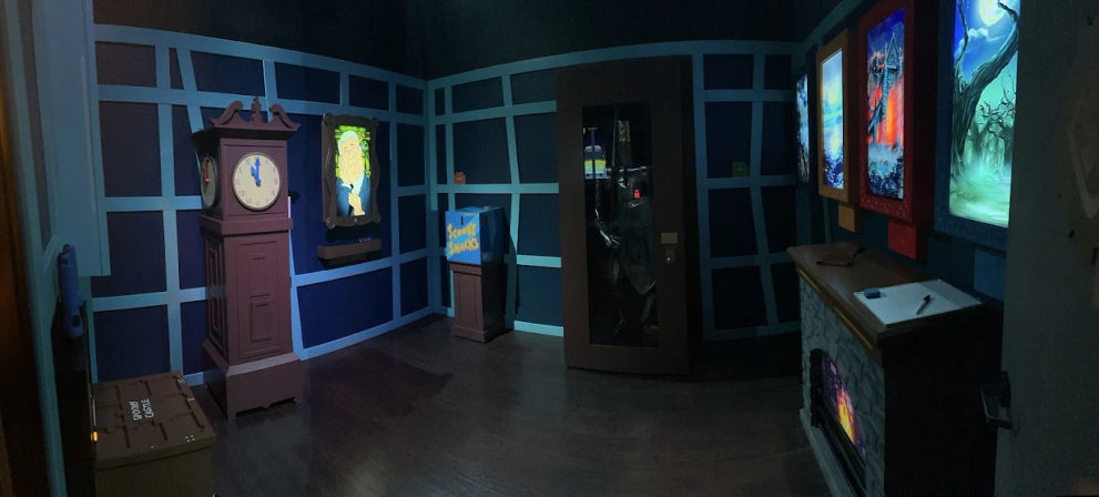 Dimly lit themed puzzle room with blue paneled walls, an antique grandfather clock, illuminated portrait, snack box on a pedestal, dark glass cabinet, and a glowing faux fireplace beneath framed fantasy artwork.