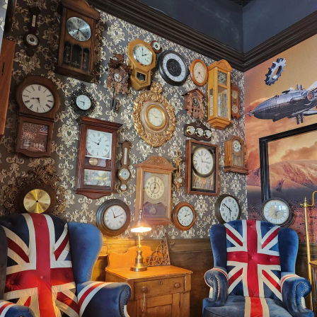 Wall filled with a collection of vintage and antique clocks above a wooden side table and two Union Jack upholstered armchairs in a cozy British‑style sitting nook.