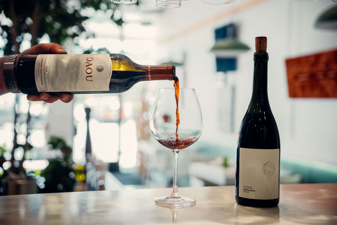 Hand pouring ruby red wine into a large stemmed glass on a marble bar counter, second bottle with cork nearby and sunlit modern bistro interior blurred in the background.