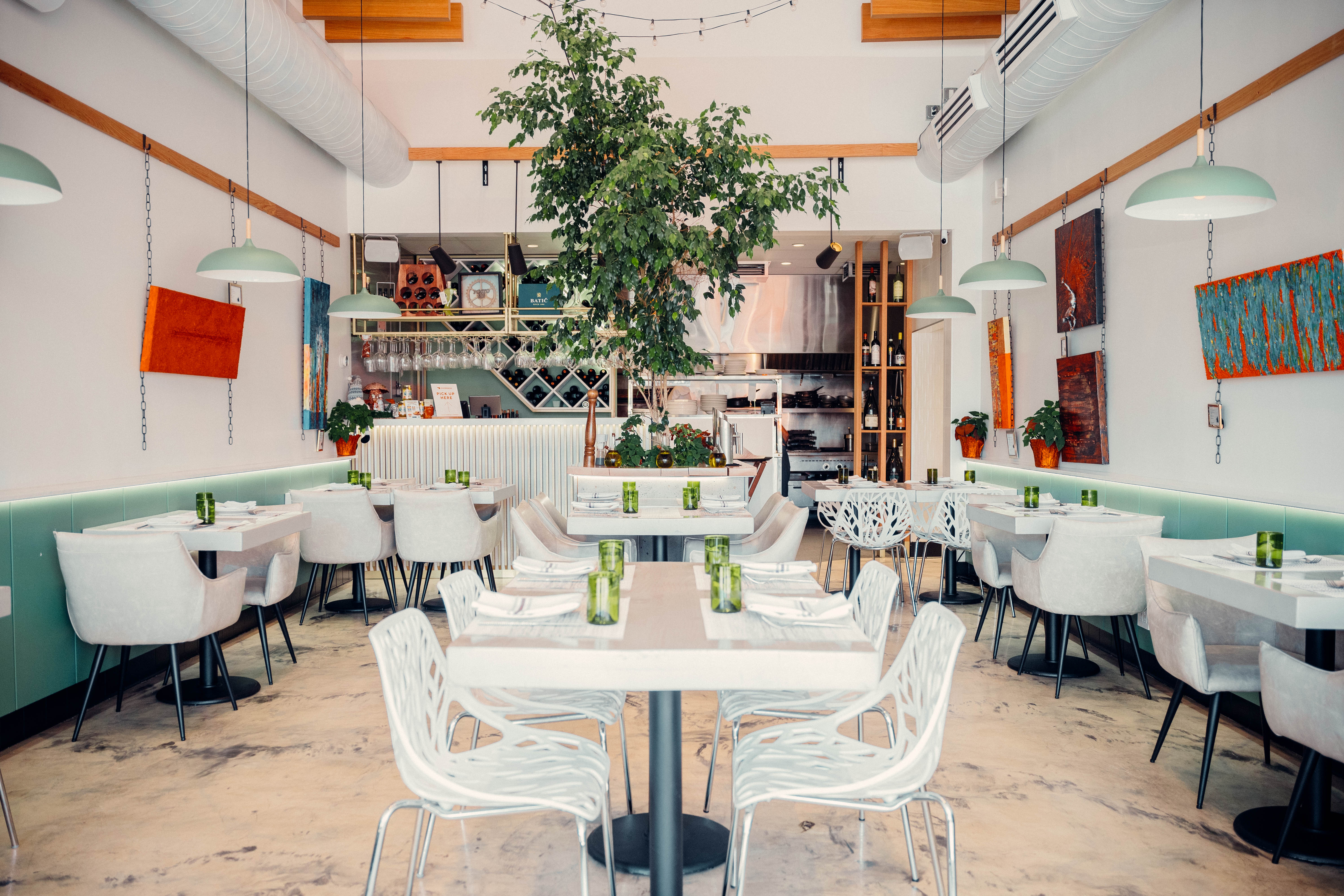 Bright modern restaurant interior with a central indoor tree, white tables and sculptural chairs, mint-green banquettes, hanging pendant lights, open kitchen and visible wine racks.