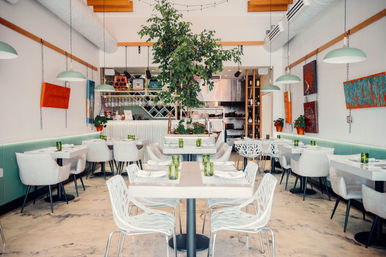 Bright modern restaurant interior with a central indoor tree, white tables and sculptural chairs, mint-green banquettes, hanging pendant lights, open kitchen and visible wine racks.