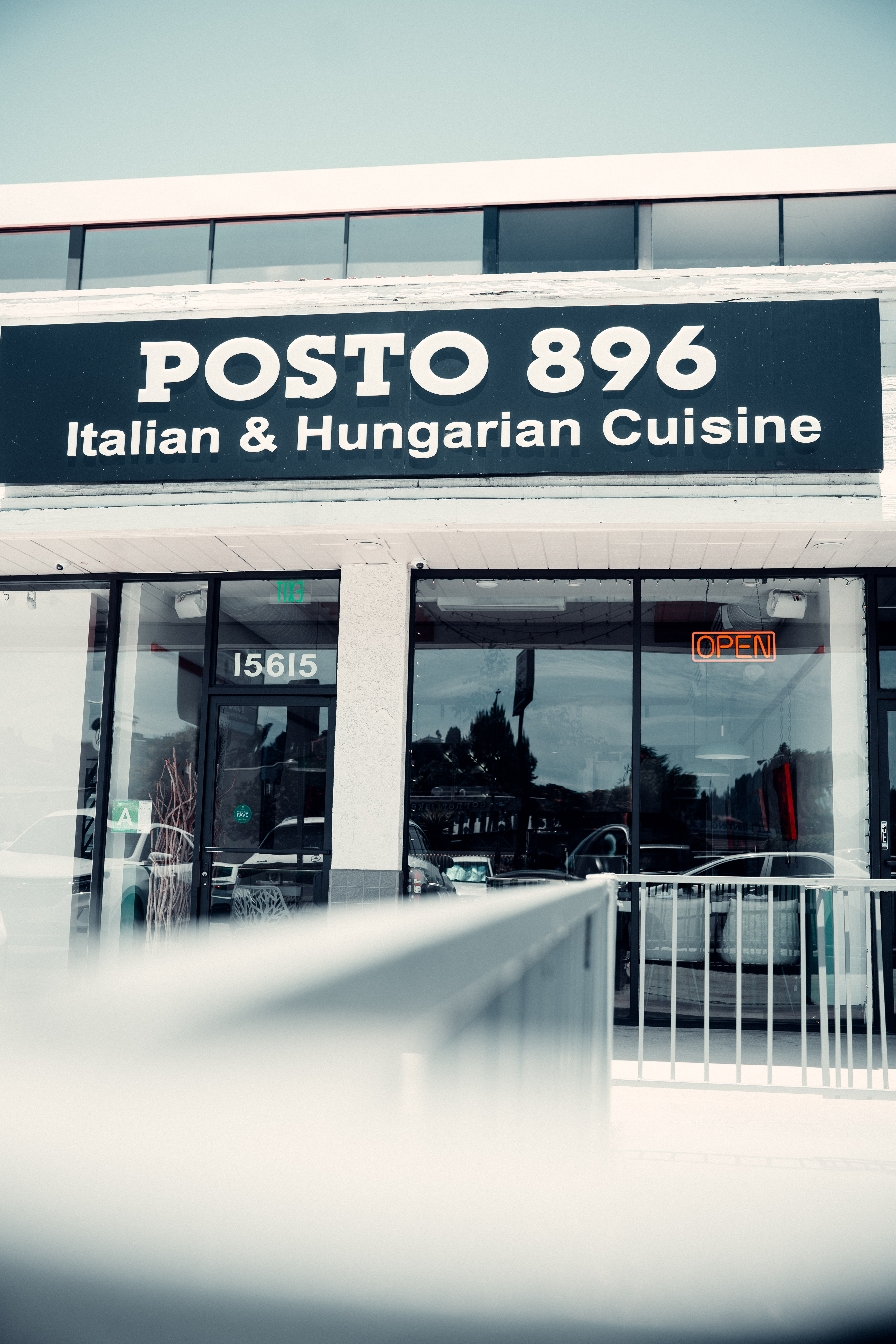 Exterior restaurant storefront with a large black sign advertising Italian and Hungarian cuisine, glass entrance doors, and an orange neon open sign.