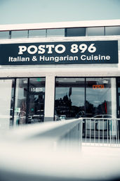 Exterior restaurant storefront with a large black sign advertising Italian and Hungarian cuisine, glass entrance doors, and an orange neon open sign.