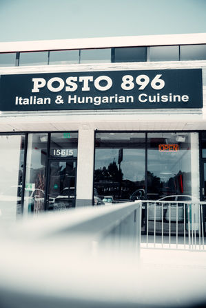 Exterior restaurant storefront with a large black sign advertising Italian and Hungarian cuisine, glass entrance doors, and an orange neon open sign.