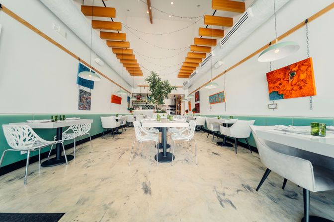 Bright modern restaurant interior with mint-green banquettes, white sculptural chairs, hanging pendant lights, wooden ceiling beams, string lights and colorful abstract art surrounding a central potted tree