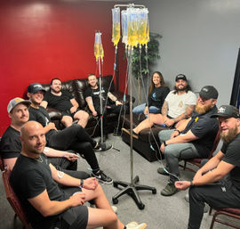 Nine adults smiling in an indoor wellness lounge seated in a circle, each connected to IV infusion lines from clear bags of yellow fluid hanging on poles
