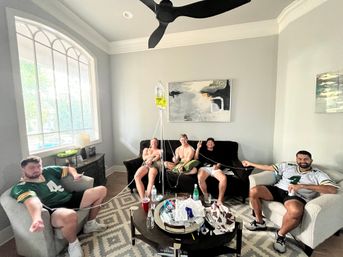 Five people lounging in a bright home living room with a large arched window and ceiling fan, wearing football jerseys and holding IV drip tubes from a hanging yellow IV bag, with a coffee table of drinks and snacks — lighthearted game-day vibe.
