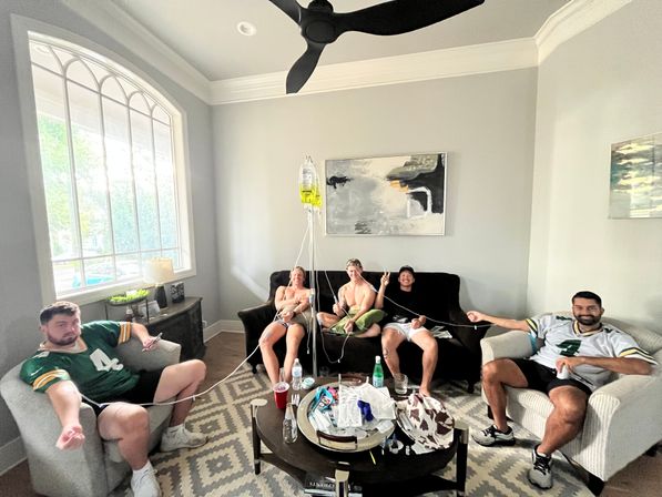 Five people lounging in a bright home living room with a large arched window and ceiling fan, wearing football jerseys and holding IV drip tubes from a hanging yellow IV bag, with a coffee table of drinks and snacks — lighthearted game-day vibe.