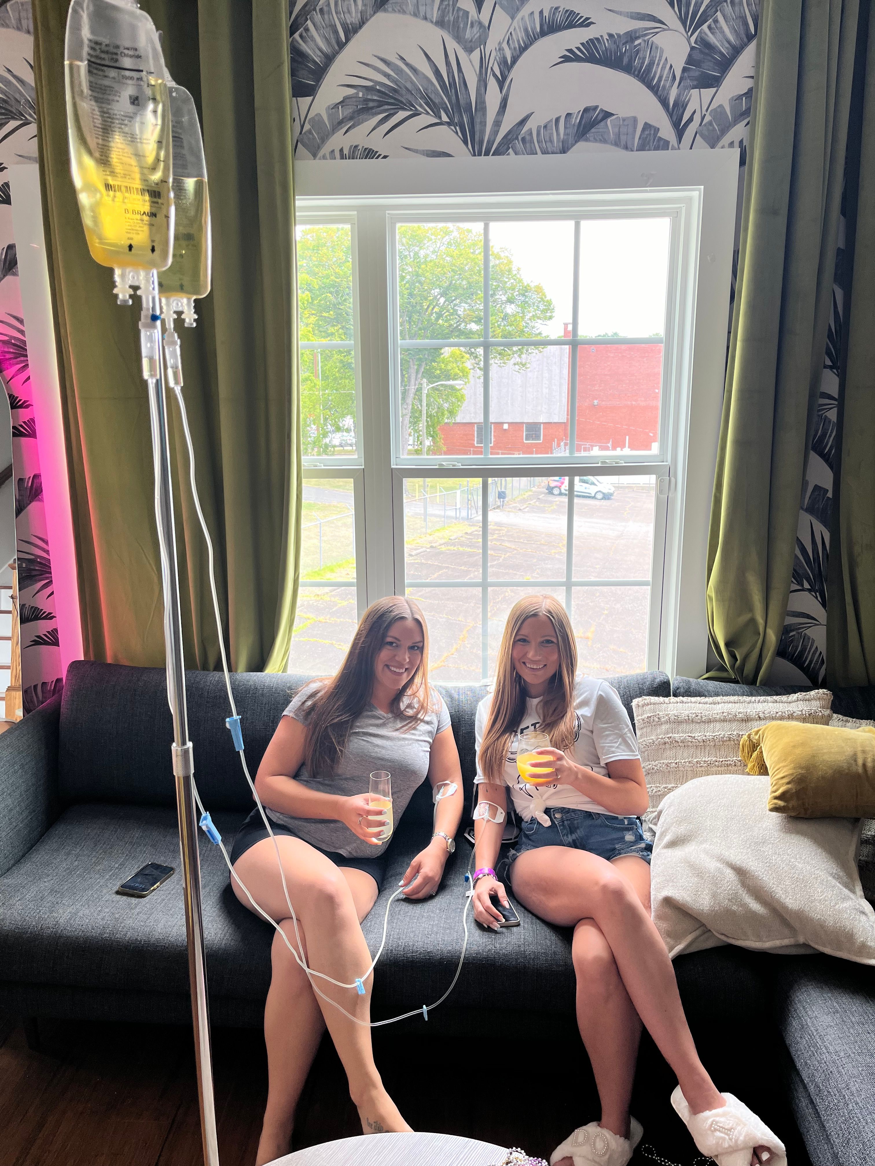 Two smiling women lounging on a gray sofa in a bright room, each receiving IV drips from a pole while holding drinks, large window with trees and a red building outside, tropical wallpaper and green curtains in the background.