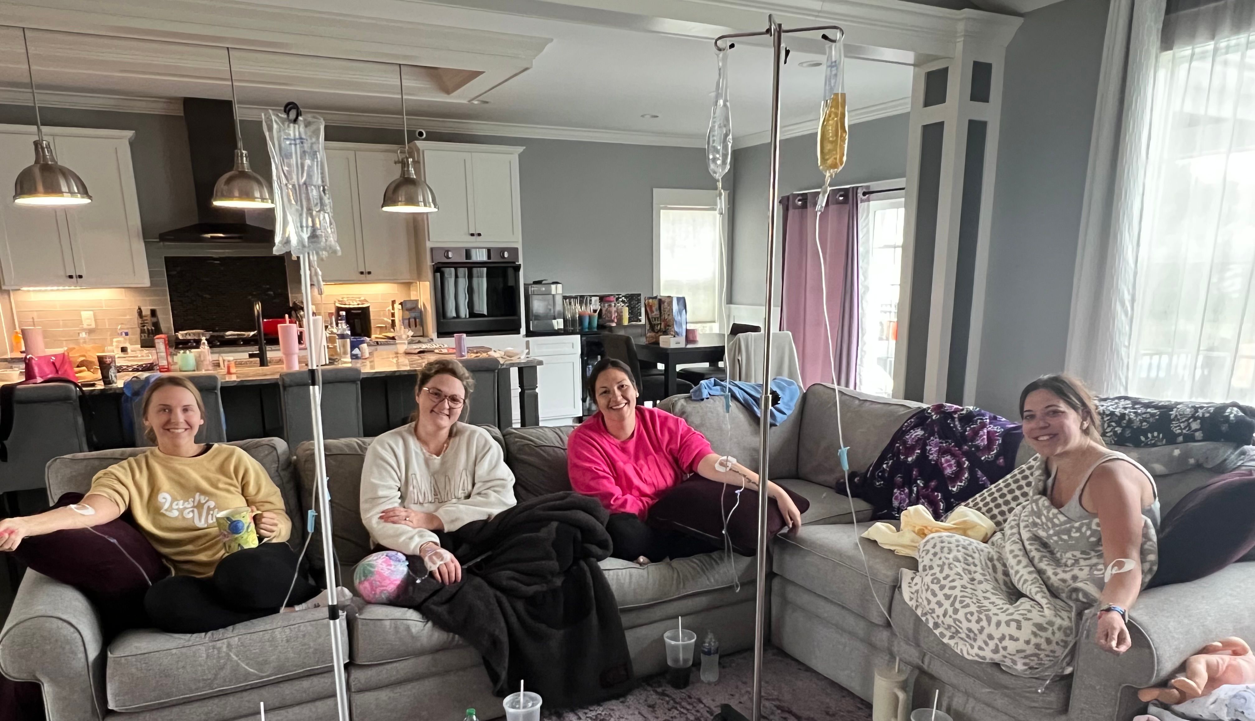 Four women relaxing on a gray sectional sofa in an open-concept living room with kitchen in the background, smiling while connected to IV bags on poles, wrapped in blankets with drinks and cozy home decor around them.