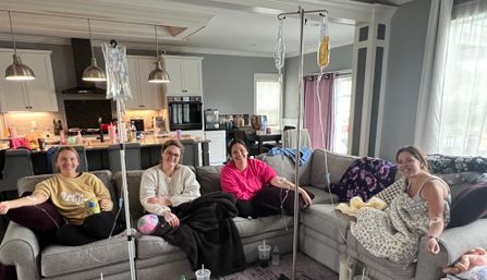 Four women relaxing on a gray sectional sofa in an open-concept living room with kitchen in the background, smiling while connected to IV bags on poles, wrapped in blankets with drinks and cozy home decor around them.