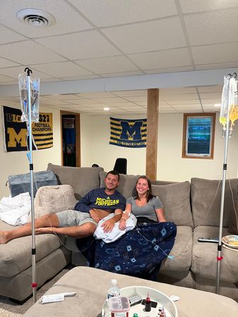 Couple smiling on a beige sectional in a basement Michigan fan cave, covered by a blue blanket and receiving IV fluids from two poles, with University of Michigan banners on the wall and a TV remote and snacks on the ottoman.