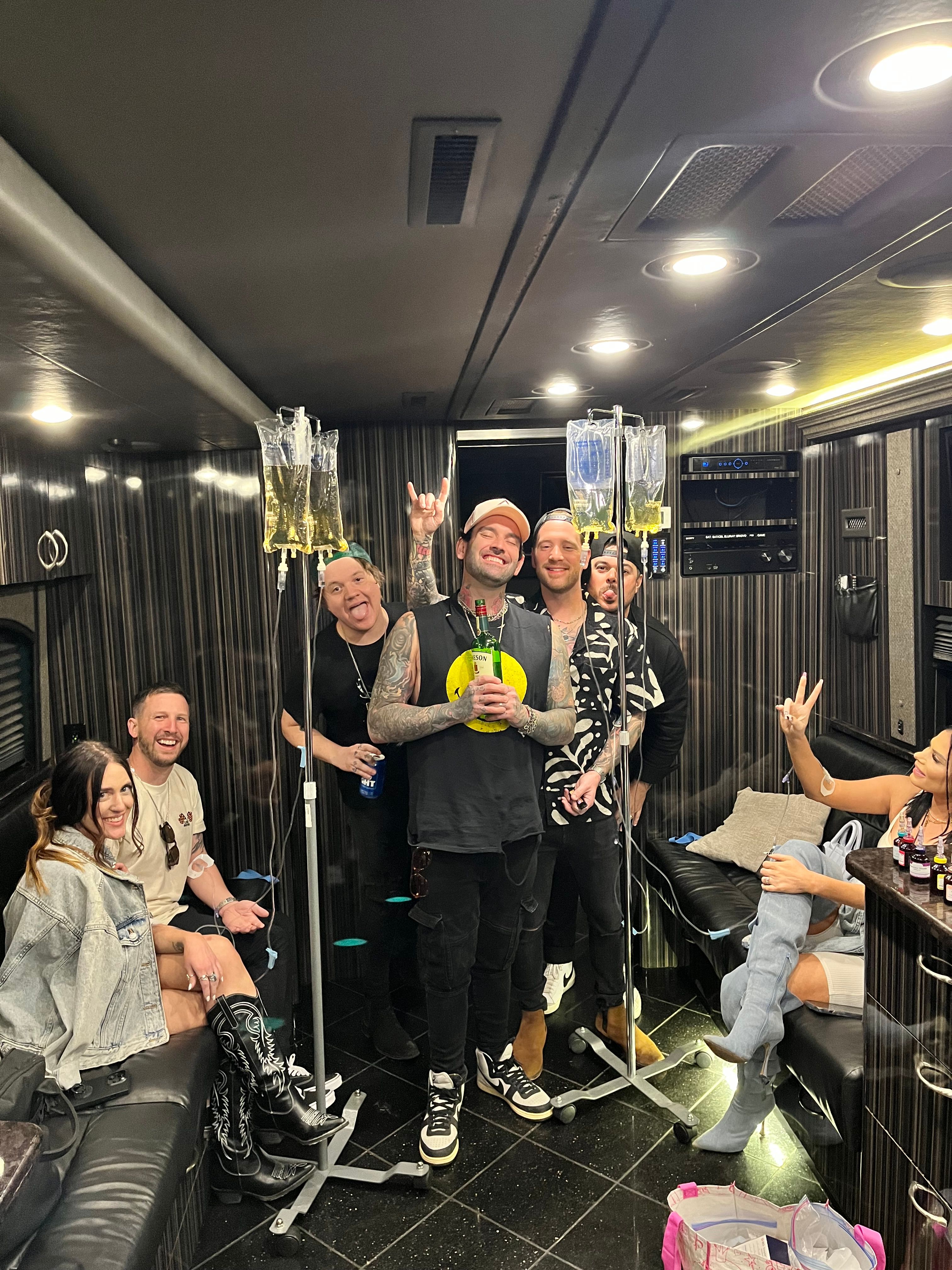 Group of friends partying inside a wood-paneled tour bus lounge, smiling and posing around IV drip stands with vitamin bags and drinks — playful on-the-road celebration scene.