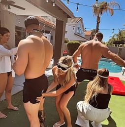Sunny suburban backyard pool party with friends wearing blindfolds playing a playful game around two shirtless men in swim trunks by a pool under string lights