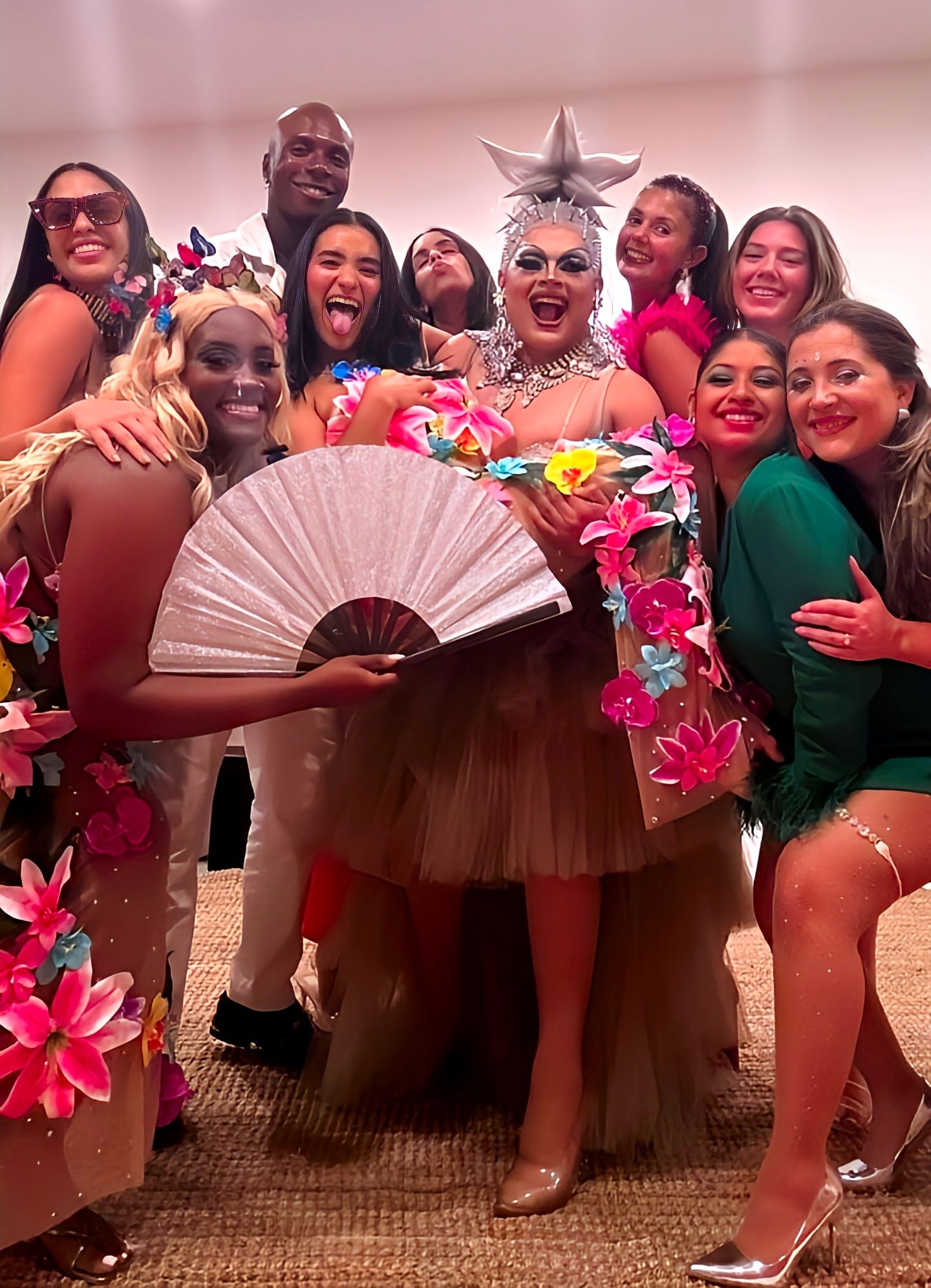 Indoor group photo at a lively drag show celebration — smiling performers and friends in colorful floral costumes, a central drag artist in a star headpiece and jeweled collar holding a large fan.