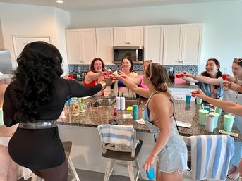 Cheerful group of women in swimwear and casual clothes gathered around a kitchen island, clinking red shot cups in a lively vacation-style toast.