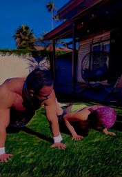 Adult and child doing push-ups on a green lawn in a sunny backyard with palm trees, patio cover and hanging egg chair — playful outdoor workout.