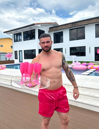 Poolside scene: shirtless man in hot-pink swim trunks holding a tray of pink plastic champagne flutes by a modern white two-story house and backyard pool with pink inflatable floats under a blue sky.