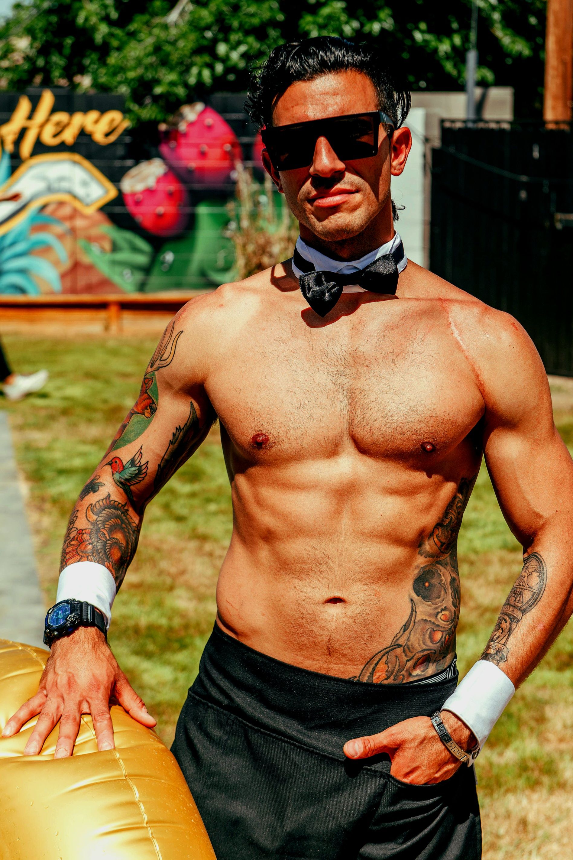 Shirtless fit man in sunglasses, black bow tie and white cuffs showing tattoos and defined abs while posing in a sunny backyard in front of a colorful strawberry mural.