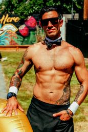 Shirtless tattooed man in sunglasses, bow tie and cuffs posing in a sunny backyard garden party, leaning on a gold inflatable with a colorful mural in the background.