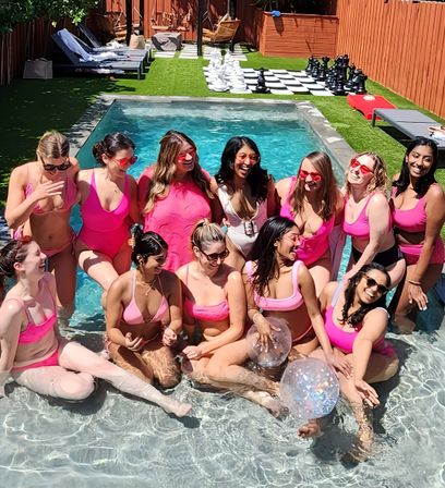 Cheerful group of women in pink swimsuits posing in the shallow end of a sunny backyard pool, laughing with clear inflatable balls, lounge chairs and a giant chessboard on turf behind them.