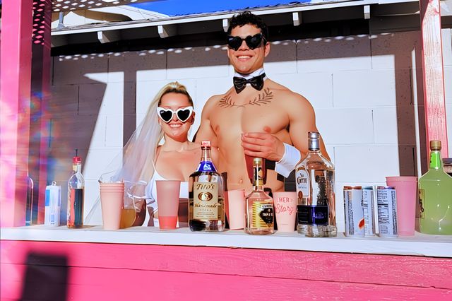 Sunlit pink poolside bar with a veiled woman in heart-shaped sunglasses and a shirtless bartender wearing a bow tie, smiling behind a counter of liquor bottles, cans and pink cups for a playful outdoor party vibe.
